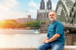 bearded man sitting on embankment of Rhine on background of Cologne Cathedral and Hohenzollern Bridge in Koel, Germany. Tourism and travel by Germany concept