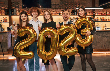 Wall Mural - With 2023 numbers balloons. Group of people in beautiful elegant clothes are celebrating New Year indoors together