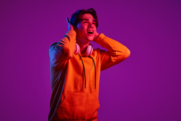 Wall Mural - Portrait of young man in casual hoodie cheerfully posing with headphones isolated over purple background in neon light