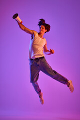 Wall Mural - Portrait of young man in white T-shirt, singlet posing in a jump over purple background in neon light