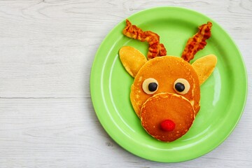 Wall Mural - Reindeer pancake made it from pancakes,bacon,cheeses,black olives and raspberry on green plate with white wood background.Creative art food idea for kids Christmas breakfast.Top view.Copy space