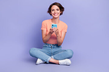 Full length photo of adorable young woman sit floor typing hold device dressed stylish striped clothes isolated on purple color background