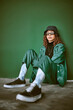 © C D/peopleimages.com - Fashion, style and clothes with a model black woman sitting on the floor against a green background. Portrait, glasses and fashionable with a young female posing to promote trendy or edgy clothing