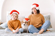 Merry Christmas and Happy New Year. Happy family with dog jack russell are waiting for new year in santa claus hats, sitting side by side on bed at home in bedroom. Mom and happy daughter in sweaters
