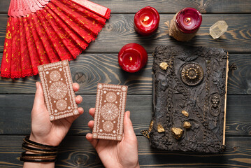 Wall Mural - Tarot cards in fortune teller hands and magic book on the old wooden table background top view.