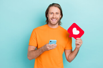 Wall Mural - Photo of cheerful positive guy wear orange t-shirt rising heart like sign typing modern device isolated teal color background