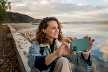 Wall Mural - Young cheerful smiling woman traveler in a denim jacket taking a picture on a smartphone on the shore of a lake in autumn weather