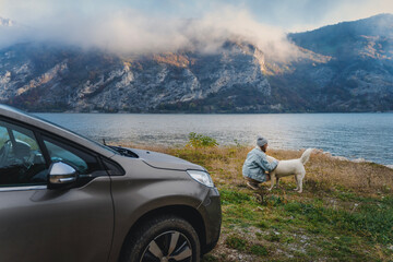 Young woman traveler standing next to the car while traveling with her white swiss shepherd dog on the shore of a mountain lake