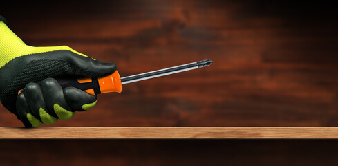 Manual worker with black and green protective work gloves holding a phillips head screwdriver ( cross shape) with black and orange plastic handle on a wooden workbench with copy space.