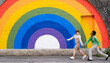 © VISTA by Westend61 - Smiling lesbian couple holding hands running on footpath in front of rainbow colored wall