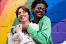 Happy Woman Embracing Girlfriend In Front Of Rainbow Flag Painted On Wall