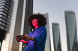 © VISTA by Westend61 - Red light falling on woman face wearing smart glasses holding smart phone in front of building