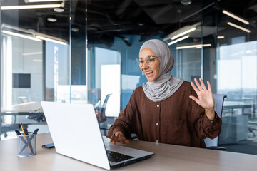 Wall Mural - Businesswoman in hijab works inside a modern office building, Muslim talks on video call, online meeting with colleagues and clients, woman cheerfully waves her hand and greets interlocutors online.