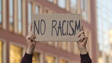 NO RACISM On Cardboard Poster In Hands Of Male Protester Activist. Stop Racism Concept, No Racism. Rallies Against Racism And Police Brutality. Peaceful Life Of Blacks Matters. Close Up.