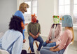 © Studio Romantic - Absurdity. Conversation of young people in rubber masks on their heads during support group meeting. Unknown man in dinosaur mask talks to people who also wear animal heads and sit in circle on chairs