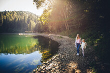 Mom And Daughter Walk Hand In Hand Along The Shore Of A Mountain Lake On A Bright Sunny Day. Coniferous Forest Around.