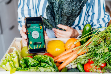 Wall Mural - No face woman holding phone with active online mobile app of Veganuary diet program and fresh kale above wooden box with vegetables. Healthy eating, weight loss. Food delivery, recipe box.