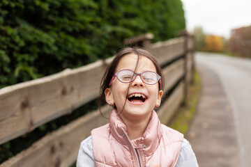 Wall Mural - Little beautiful cute girl in glasses laughing near a wooden fence. Village child in a vest on a walk in the countryside