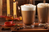 Fototapeta Kwiaty - Two transparent glasses with cappuccino with black coffee, milk foam and cinnamon in autumn setting, burning candles, pumpkins, yellow and red tree leaves on wooden background