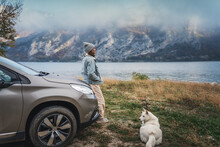 Young Woman Traveler Standing Next To The Car While Traveling With Her White Swiss Shepherd Dog On The Shore Of A Mountain Lake