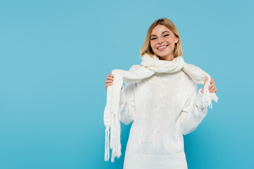 Sticker - happy blonde woman in white sweater adjusting winter scarf isolated on blue.