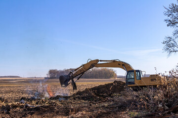 Close up view of a heavy equipment excavator moving trees and wooden debris into a fire pit near the area of a demolished old farm building