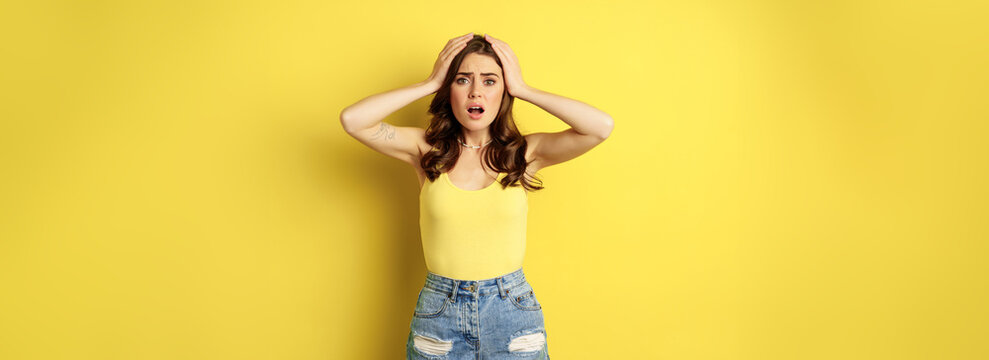Troubled and shocked woman holding hands on head, panicking, feeling distressed and anxious, having problem, standing over yellow background