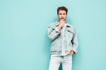 Portrait of handsome confident stylish hipster lambersexual model with curly hairstyle. Sexy man dressed in jeans jacket. Fashion male isolated on blue wall in studio