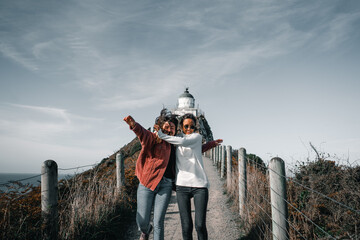 Wall Mural - two caucasian girls jumping hugging having fun happy smiling running down the narrow dirt road leading to the white lighthouse, nugget point, new zealand