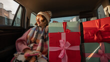 Young Woman Dressed For The Holidays Drives In Car On Back Seat With Beautifully Wrapped Christmas Presents. In Anticipation Of The Winter Holidays And Shopping. Wide Angle Shot