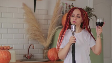 Wall Mural - Young woman with bright hairstyle with retro microphone and drinking red wine, dancing in kitchen. Female singer with dreadlocks singing into microphone entertaining with glass of wine at home.