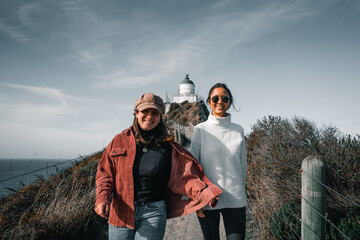 Canvas Print - two young caucasian women walking smiling happily down the narrow dirt road coming from the lighthouse next to the wooden fence, nugget point, new zealand