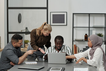 Poster - Group of intercultural displeased employees bullying African American male colleague sitting by workplace in front of laptop