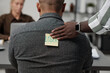 © pressmaster - Hand of black man putting sticker with written word loser on back of colleague while passing by towards his workplace in office