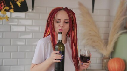 Wall Mural - Young woman with bright dreadlocks drinking red wine, dancing in kitchen. Portrait of adult female entertaining with glass and bottle of wine at home.