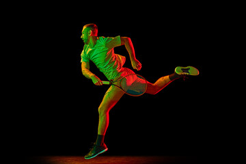 Dynamic portrait of energetic male tennis player in sportwear playing tennis isolated over dark background in neon light. Concept of motion, speed, professional sport.