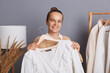 Indoor shot of Caucasian happy beautiful woman holding clothes on racks in modern fashion store, showing white shirt from a new collection, consultant looking at camera with toothy smile.