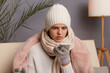 Portrait of shocked scared woman sitting on the sofa at home wearing winter coat, hat and mittens, holding cell phone, looking at display with big eyes.