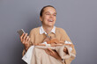 Portrait of joyful cheerful Caucasian woman standing with clothing in hands and holding mobile phone , laughing happily, enjoying shopping, posing with new attires isolated over gray background.
