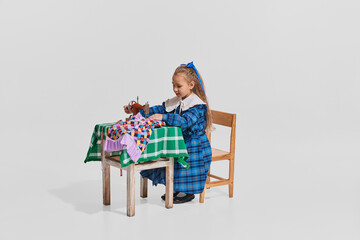 Portrait of pretty little girl in cute blue dress sitting at the table and sewing isolated over grey background