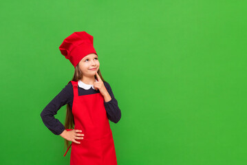 A little chef girl in an apron and a cooking cap points to your advertisement on a green isolated background. Copy space.