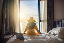 Tourist Woman Sitting On Bed Nearly Window, Looking To Beautiful Sunset Above Sea Or Ocean, Reflection Of Sun In The Water After Video Conference Meeting With Colleagues , Online Working On Laptop.  
