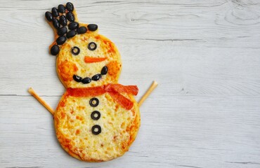 Wall Mural - Snowman pizza made it from pizza crust,pizza sauce,black olives,mozzarella cheeses,pepperoni and carrot on white wood background.Creative art food idea for celebrate Christmas.Top view.Copy space