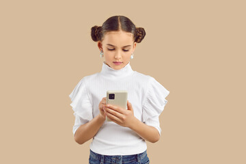 Child using modern smartphone. Little kid texting on mobile phone. Beautiful girl standing on beige studio background, searching information on Internet, reading messages, scrolling social media feed