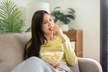 Wall Mural - Young asian woman holding a bowl of popcorn to eating popcorn while lying on the big comfortable sofa