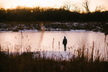 Boy Walks Out On Frozen Pond In The Winter Snow At Sunset