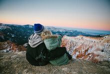 Siblings Snuggle And Overlook Snowy Canyon In Mountains At Sunset