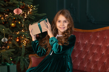 Happy little smiling girl opens a holiday gift sitting on sofa by the Christmas tree in cozy living room of the house. Beautiful girl in green dress holding christmas present in the New Year interior.