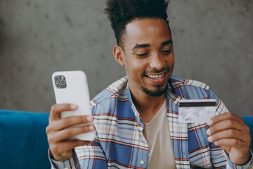 Sticker - Young fun man wear shirt sits on blue sofa couch using mobile cell phone hold credit bank card shopping online book tour stay at home hotel flat rest relax spend free spare time in living room indoor.