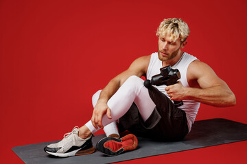 Wall Mural - Full body young strong sporty sportsman man in white clothes spend time in home gym sit on yoga mat use hand gun do muscle leg massage isolated on plain red background Workout sport fit body concept